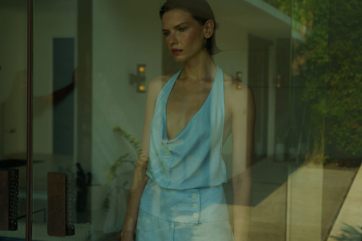 Woman standing behind a window wearing a blue dress, staring outside.