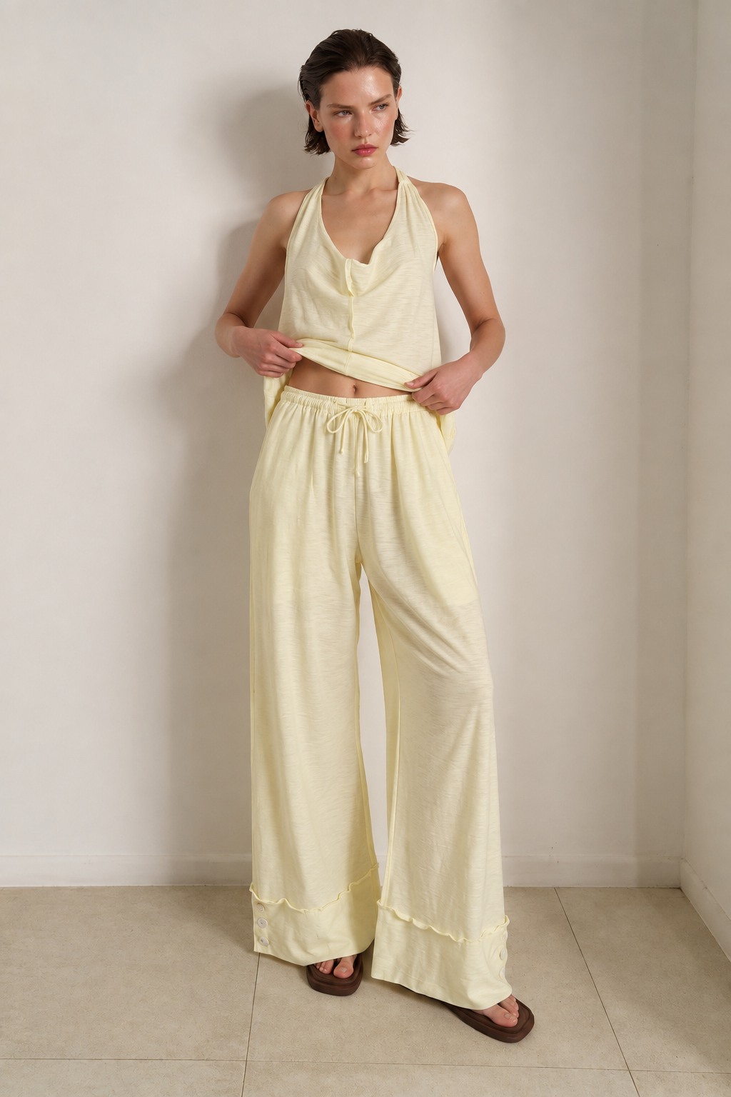 A woman modeling a pale yellow two-piece lounge set consisting of a halter-neck crop top and matching wide-leg drawstring pants, standing against a neutral beige wall.
