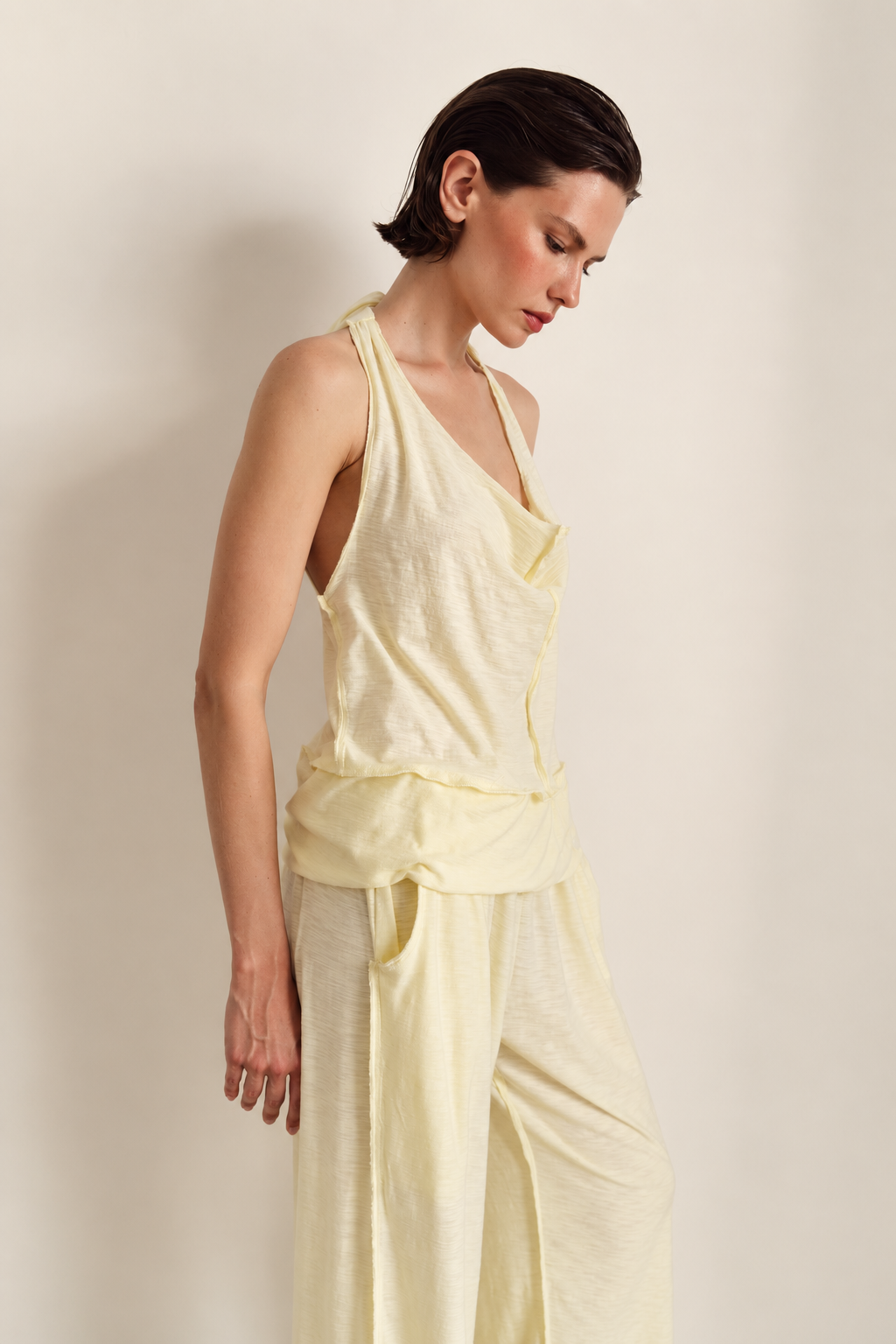 A woman modeling a pale yellow linen two-piece outfit consisting of a halter-style wrap top and matching wide-leg pants, set against a plain beige studio background.