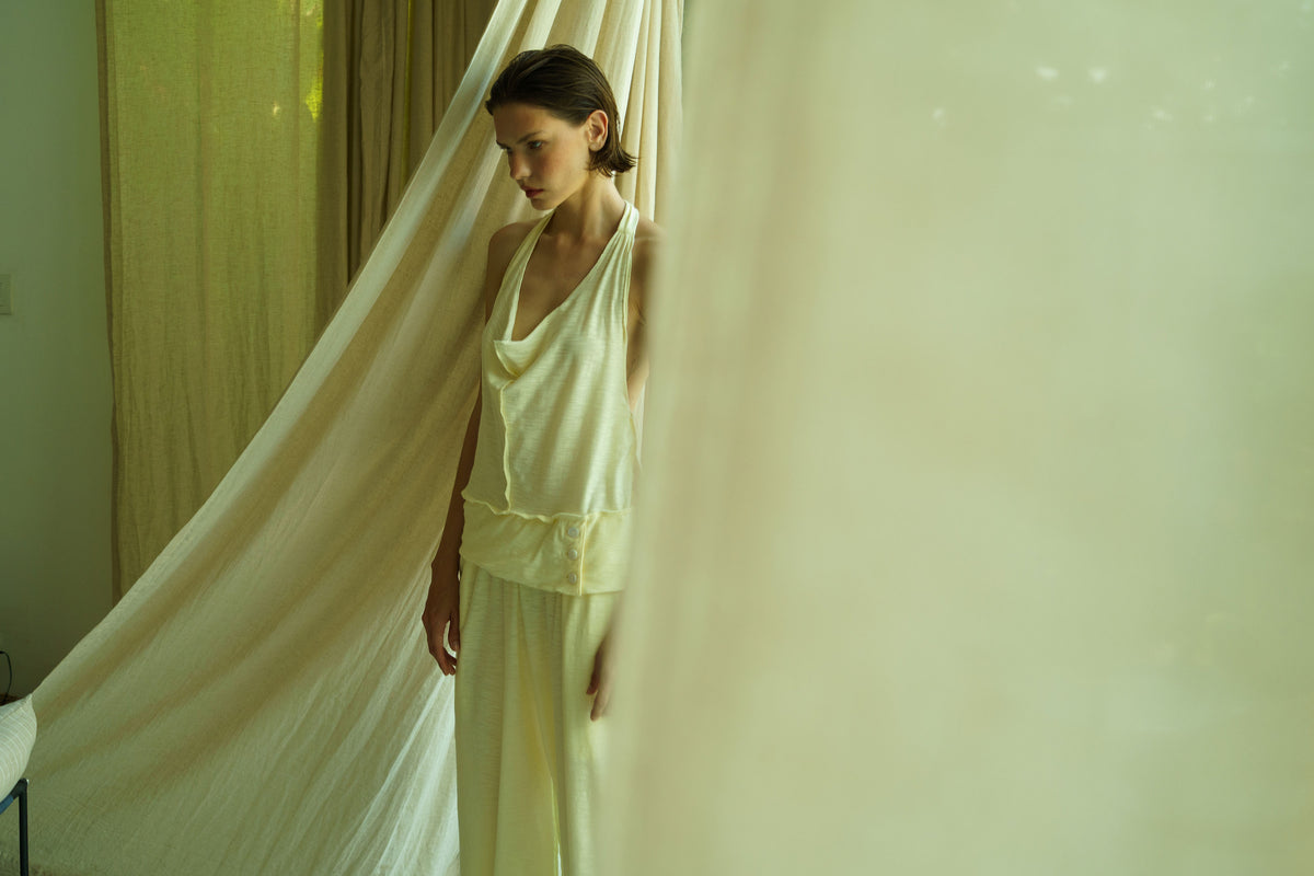 A woman wearing a pale cream sleeveless top and matching wide-leg pants stands partially hidden behind a flowing sheer curtain in a softly lit indoor setting.