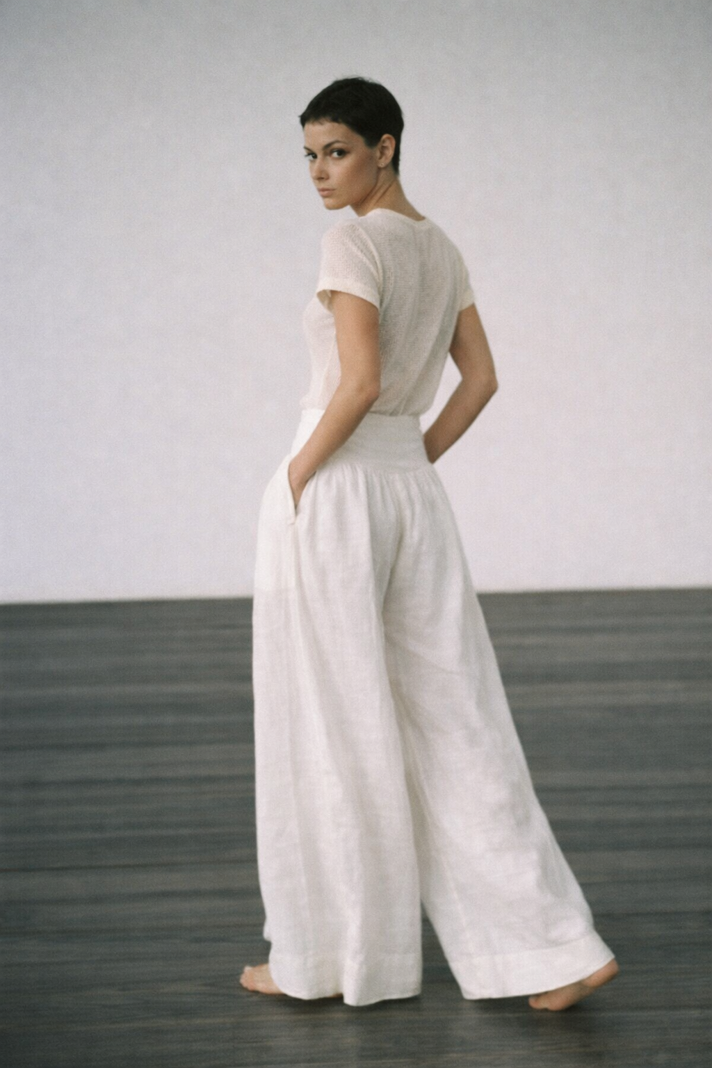 A woman stands barefoot in a minimalist studio, wearing a white short-sleeve top and matching wide-leg trousers, photographed from the back against a light gray wall and dark wood floor.
