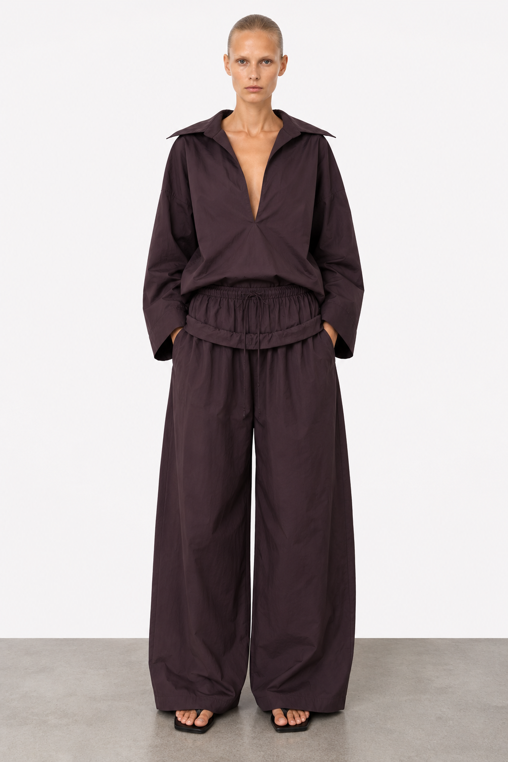 A model stands against a light gray studio background wearing a dark plum-brown matching lounge set consisting of a long-sleeve collared top and wide-leg pants.