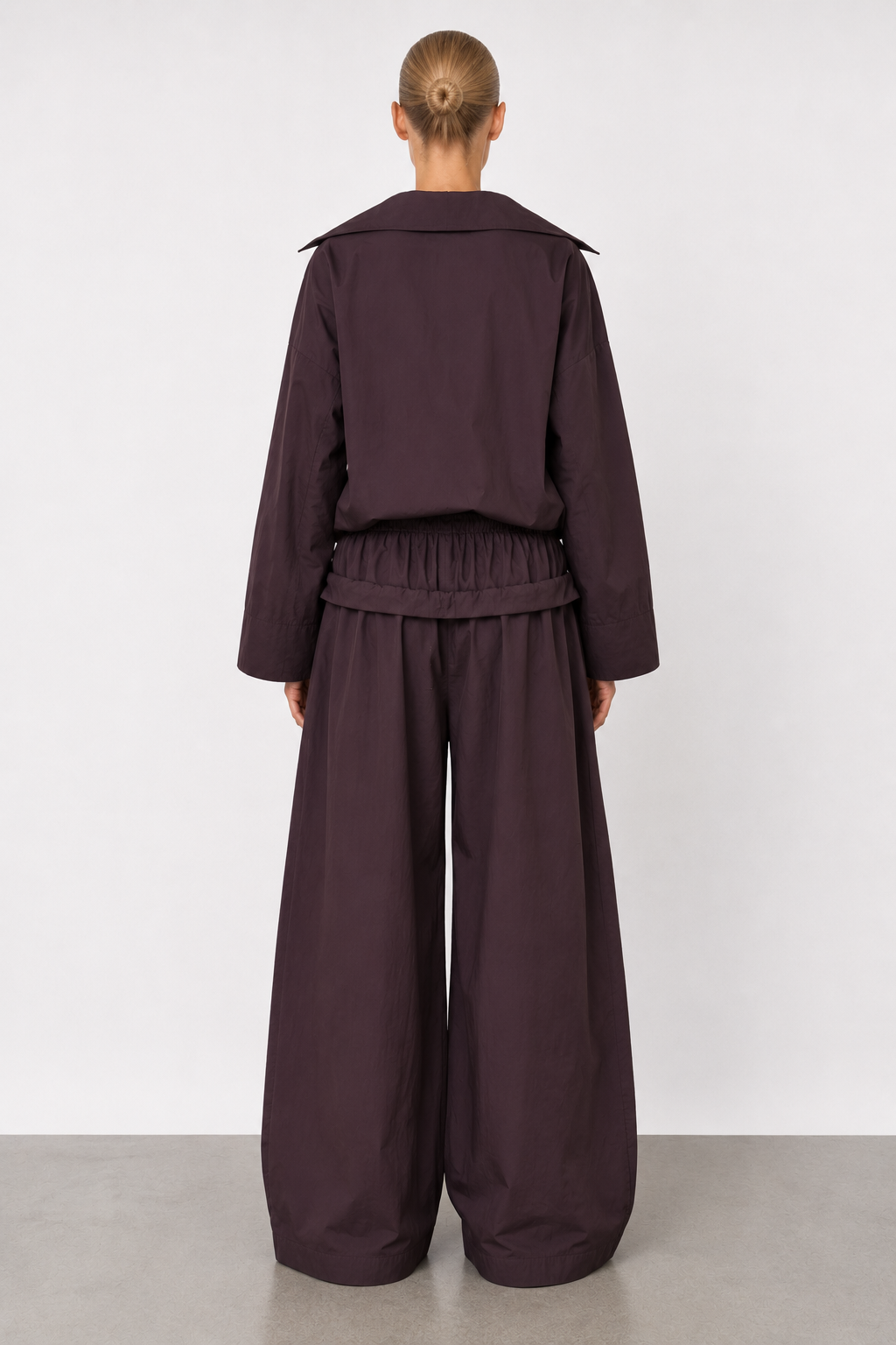 A model viewed from the back wearing a dark plum purple two-piece outfit consisting of a cropped long-sleeve top with wide shoulders and matching wide-leg trousers, set against a plain light gray studio background.
