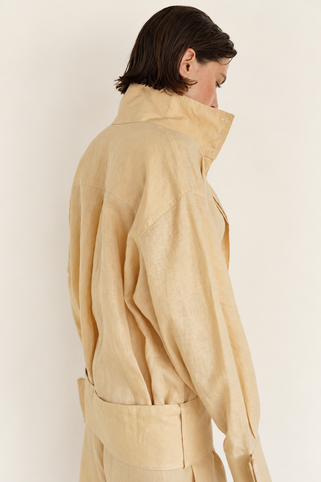 A model shown from the back wearing a light beige linen jacket with a high collar and matching trousers against a plain off-white background.