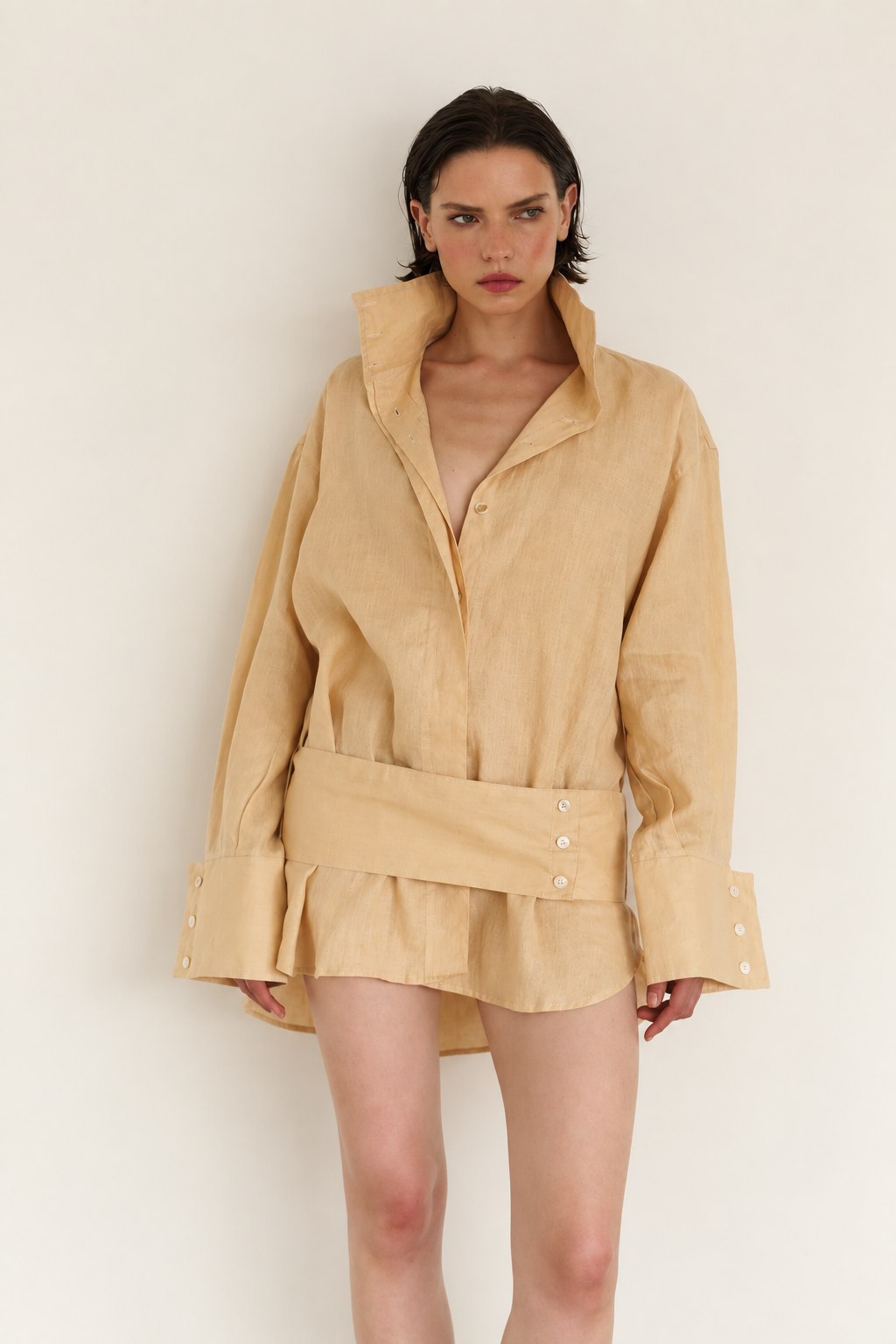A model wearing a light beige linen shirt jacket with a wide waist belt and button details, set against a plain off-white background.