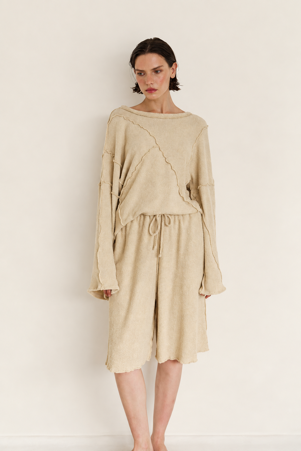 A model wearing a beige knit two-piece outfit consisting of an asymmetrical long-sleeve top and matching drawstring shorts, standing against a plain off-white wall.