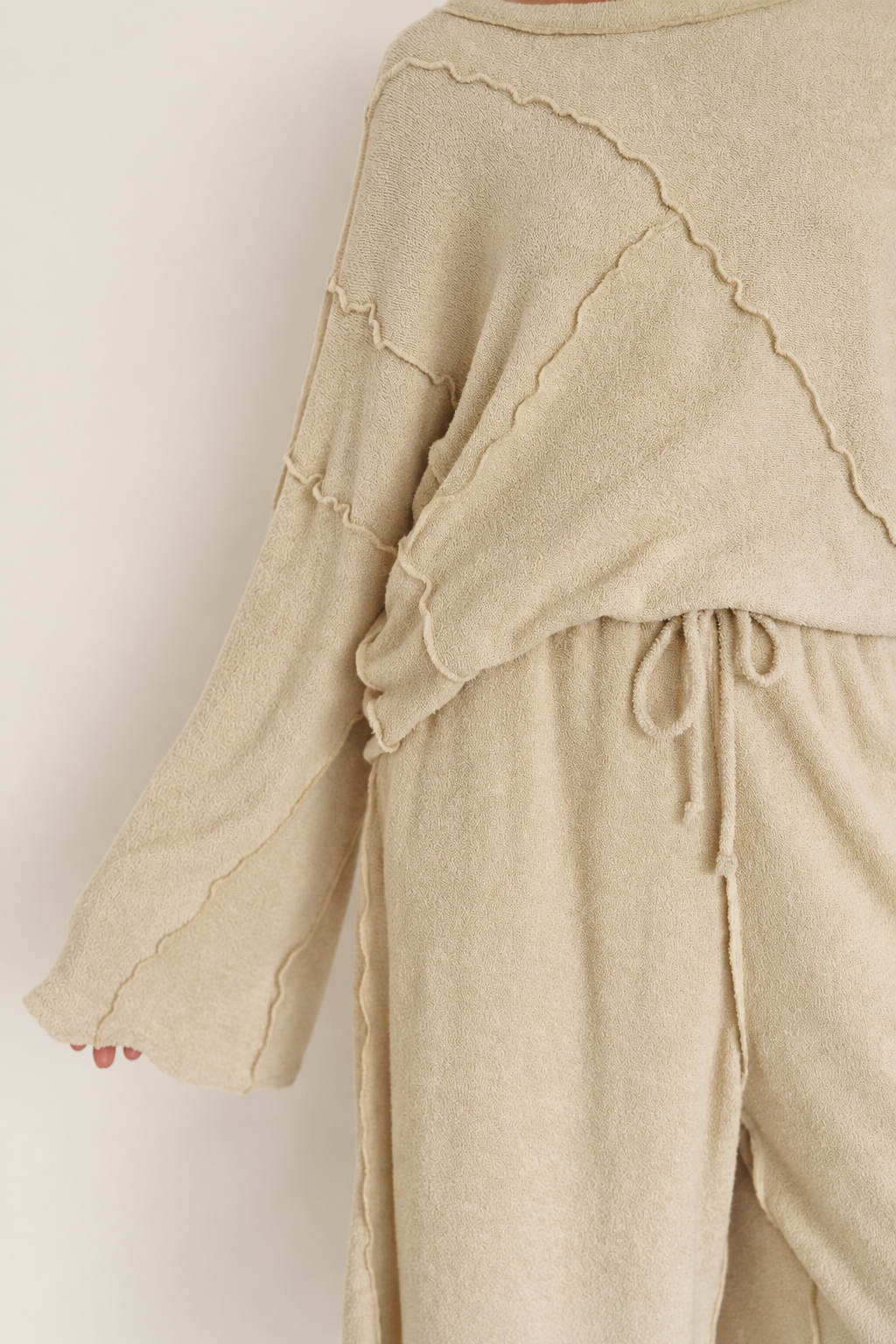 Close-up of a person wearing a soft knit beige jumpsuit or romper with long sleeves, scalloped seam details, and a drawstring waist, against a plain light background.