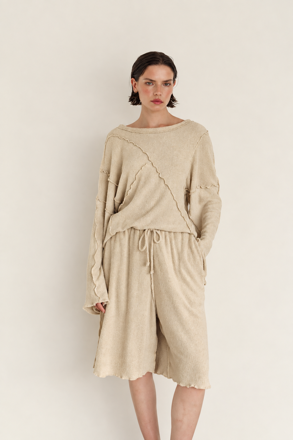 A model wearing a beige knit two-piece set consisting of a long-sleeve cropped top with diagonal patchwork details and matching drawstring shorts, set against a plain off-white background.