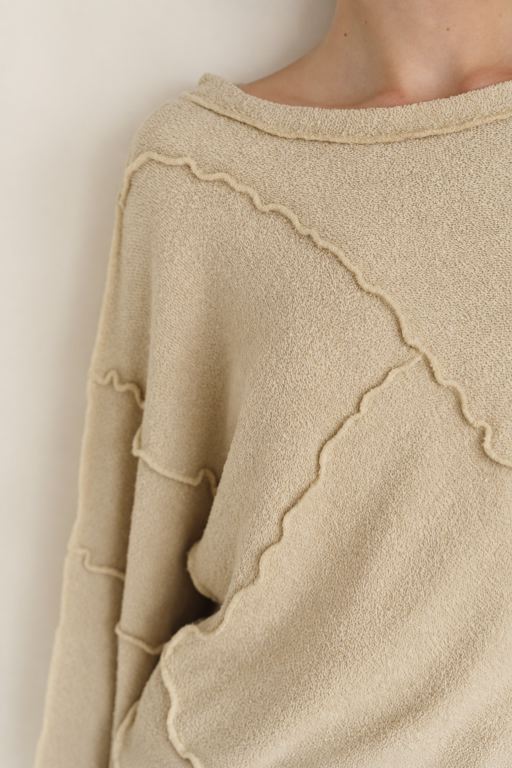 Close-up of a person wearing a soft knit sweater in a warm beige/cream tone, showing the shoulder, neckline, and part of the torso against a plain light background.