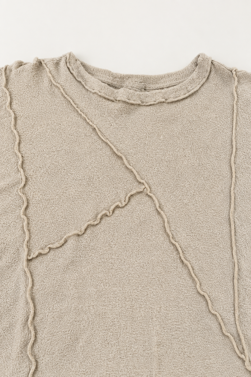 Close-up of a beige knit top with textured diagonal and vertical wavy stitched details on a plain light background.