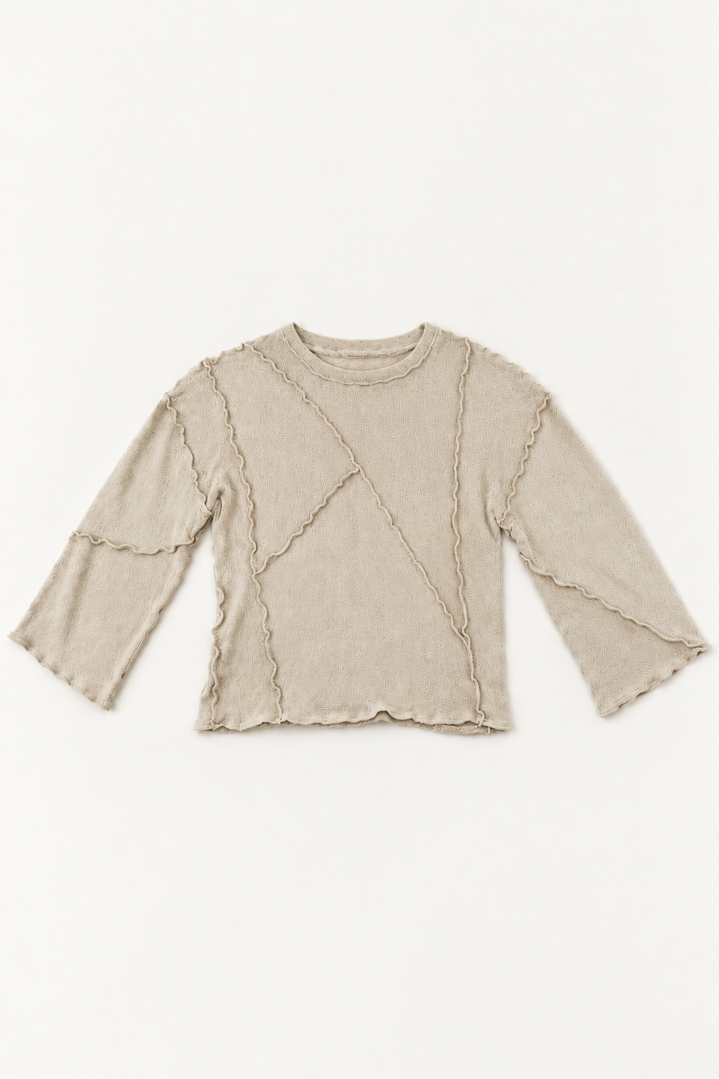 A beige knit long-sleeve crop top with asymmetrical draped details and scalloped edges, laid flat against a light off-white background.