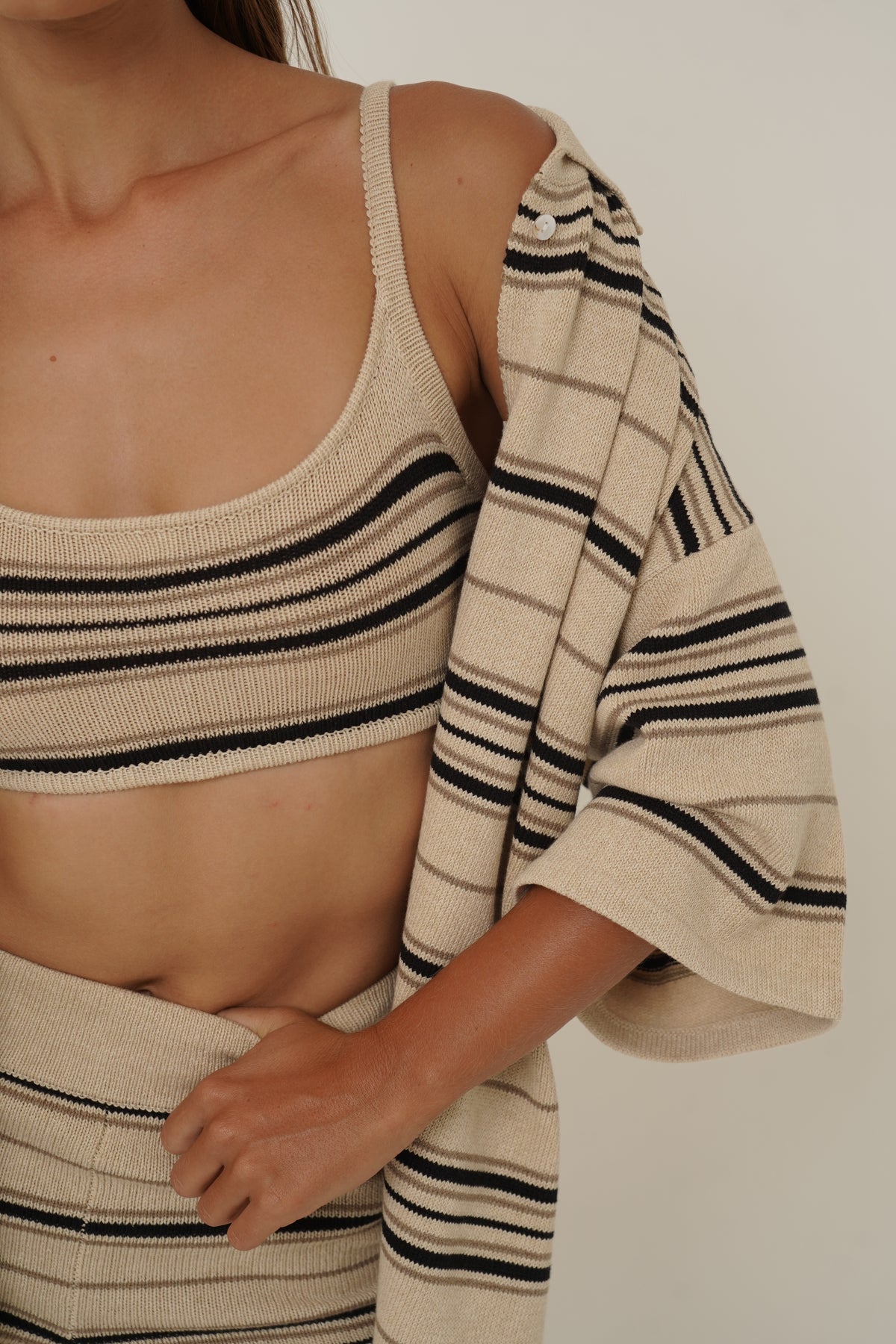 A close-up of a model wearing a beige knit top and matching striped cardigan. The top is cream-colored with thin black stripes, and the cardigan features a larger cream base with black and thin brown stripes.