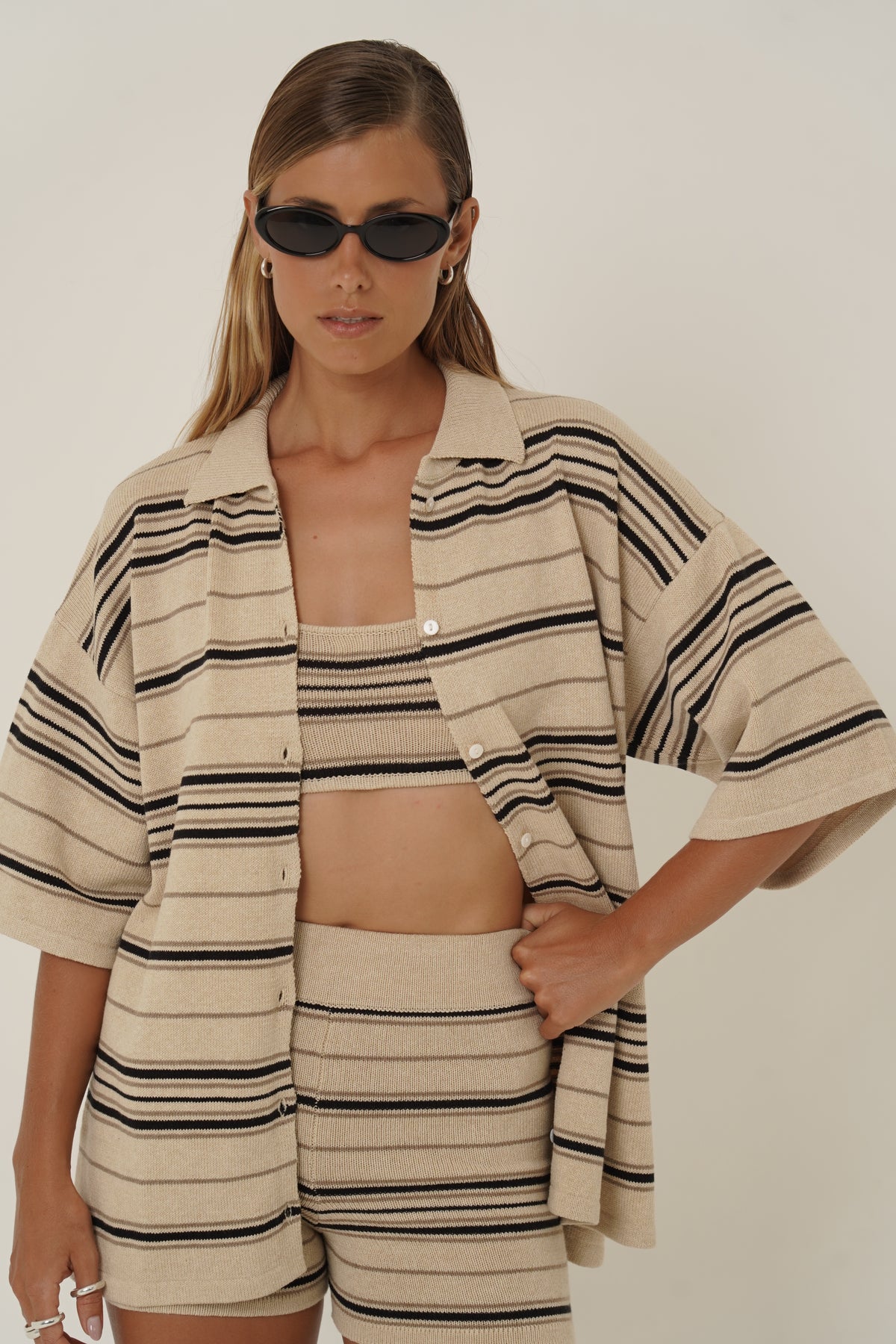 A woman wearing a beige knit matching set with black stripes, consisting of a cropped button-front cardigan and shorts, styled with black sunglasses against a plain light background.
