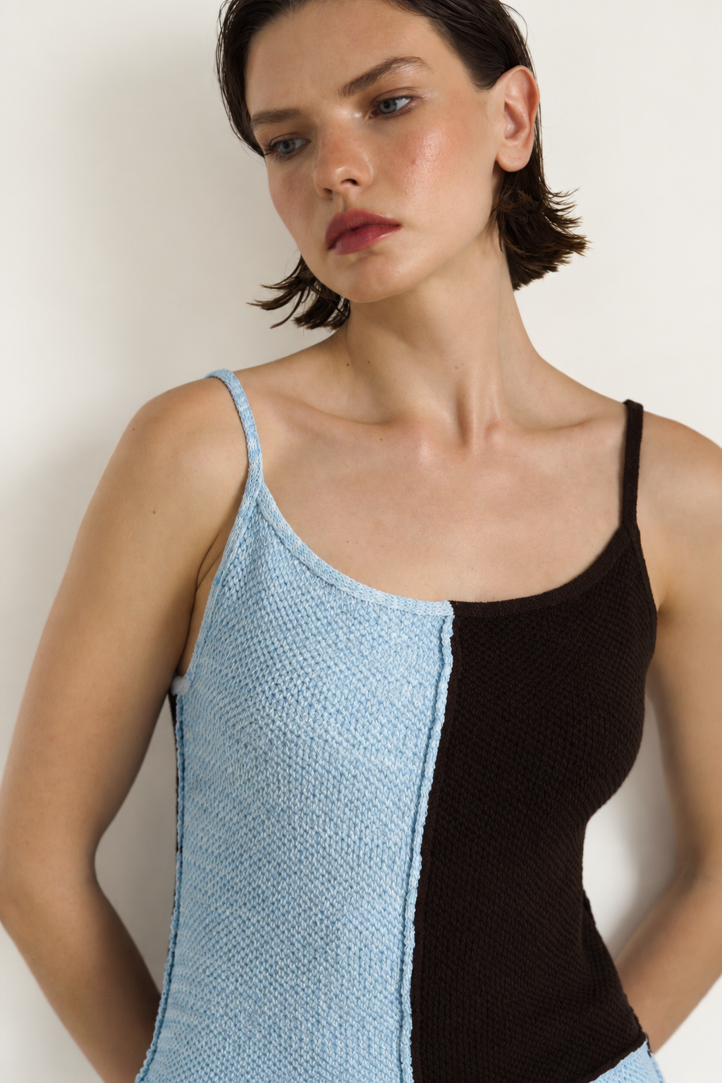 A close-up of a model wearing a color-block knit camisole with a light blue textured left side and a solid black right side, against a plain off-white background.