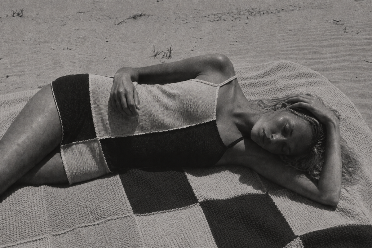 A black and white photograph of a woman lying on a beach blanket on sand, wearing a patchwork knit swimsuit.