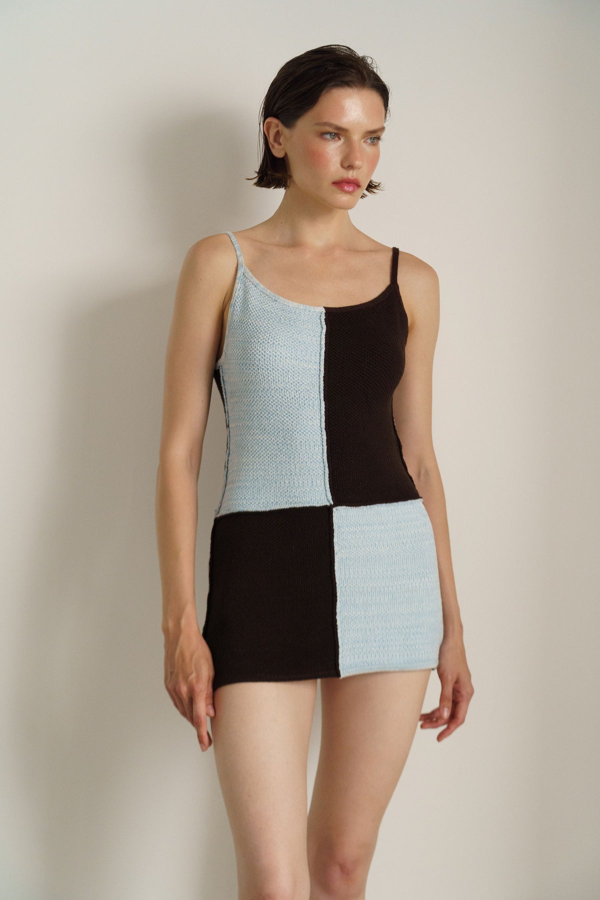 A model wearing a sleeveless mini dress with a patchwork design of light blue and dark brown textured fabric panels, set against a plain light beige background.