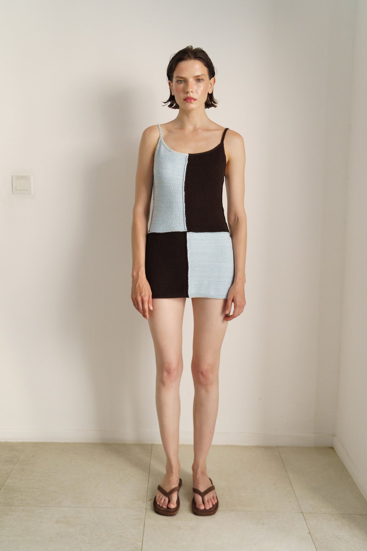 A model stands in a minimalist room wearing a color-blocked knit mini dress with thin straps, featuring a light blue, black, and white pattern, paired with brown sandals.