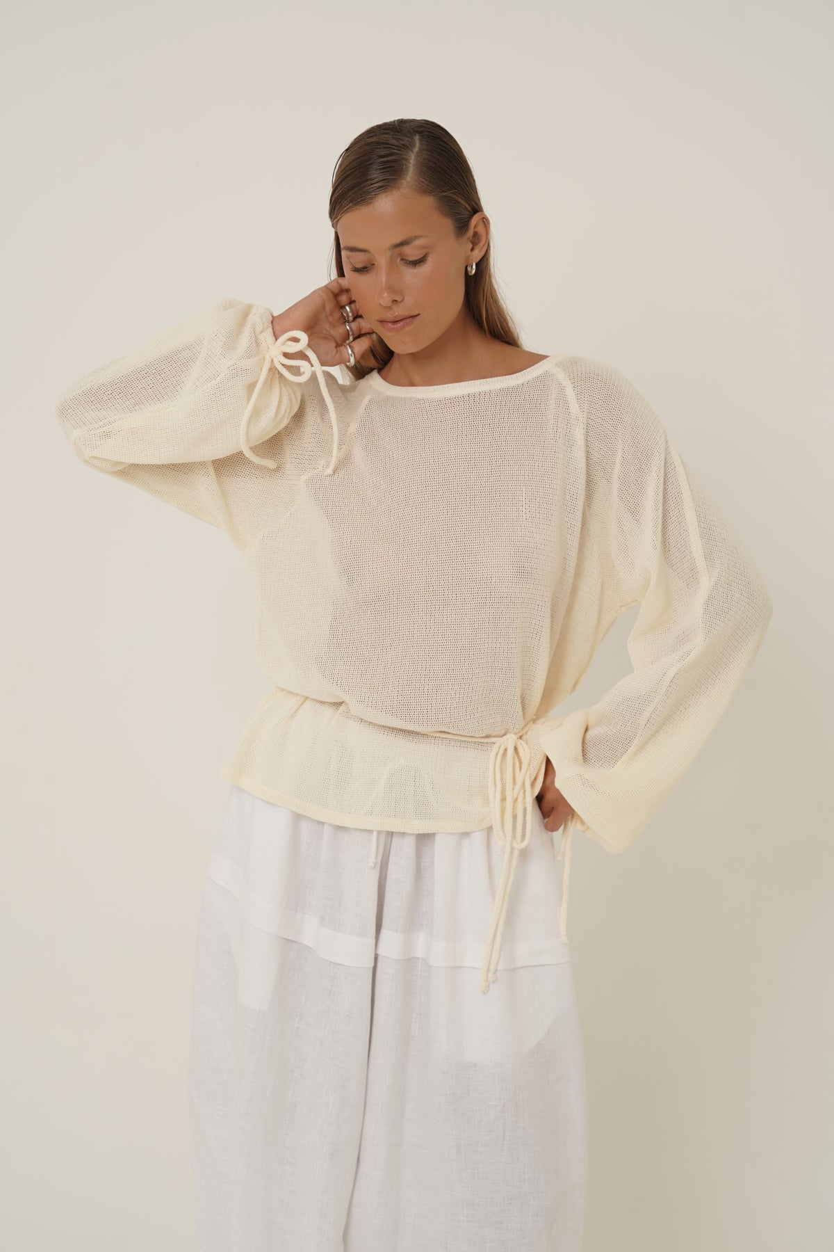 A woman wearing a cream-colored sheer long-sleeve top with tie details on the cuffs and waist, paired with white wide-leg pants, against a plain light beige background.