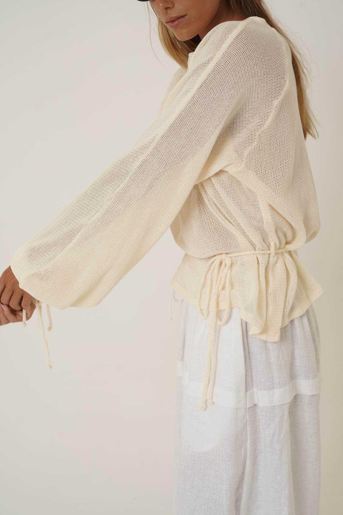 A woman wearing a cream-colored, semi-sheer long-sleeve knit top with a drawstring waist, paired with white tiered linen pants, photographed against a neutral cream background.