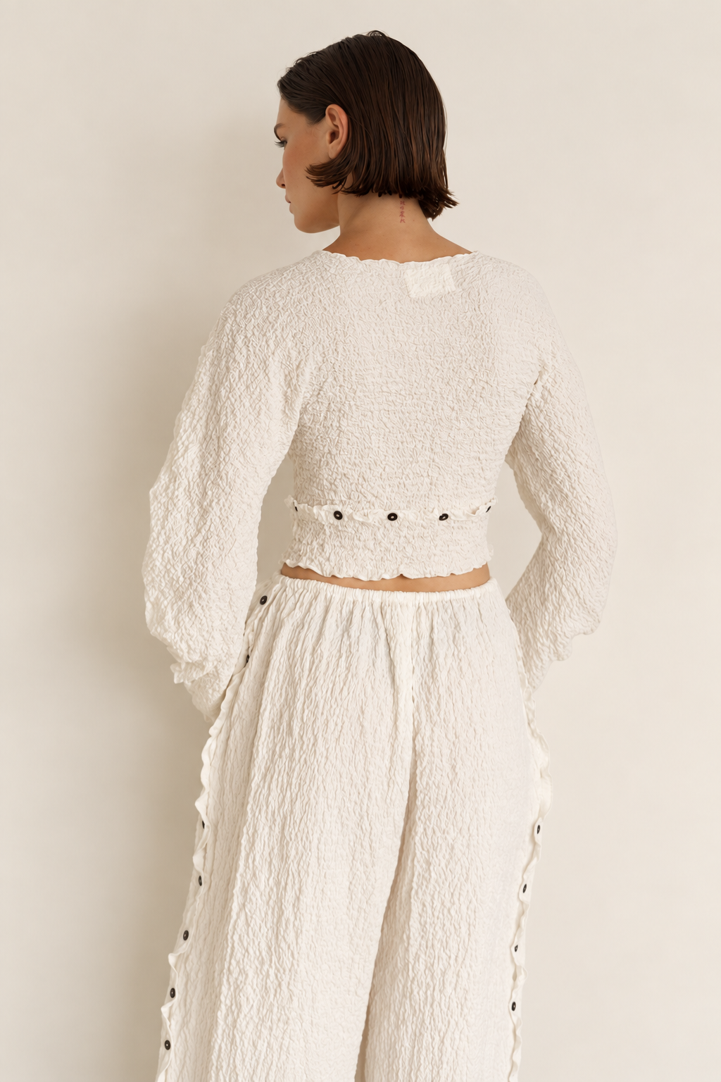 A model shown from the back wearing a textured off-white two-piece set consisting of a long-sleeve crop top with scalloped edges and button details, and matching textured wide-leg pants.