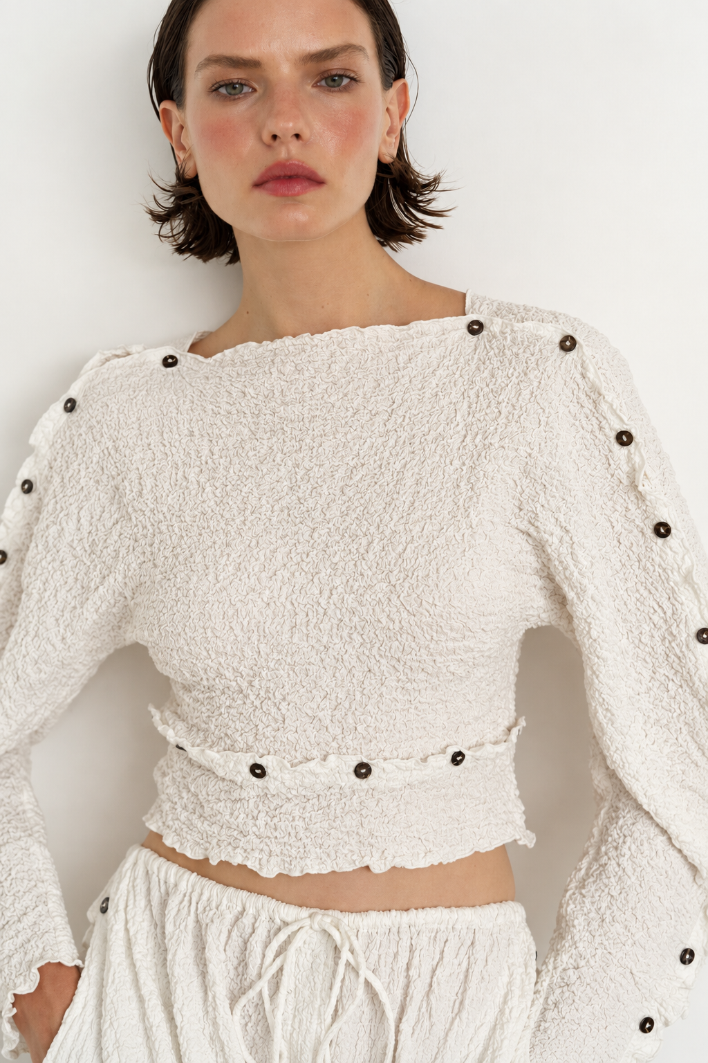 A close-up of a model wearing a textured off-white cropped long-sleeve top with dark decorative buttons along the shoulders and waist, paired with matching textured bottoms against a plain light background.