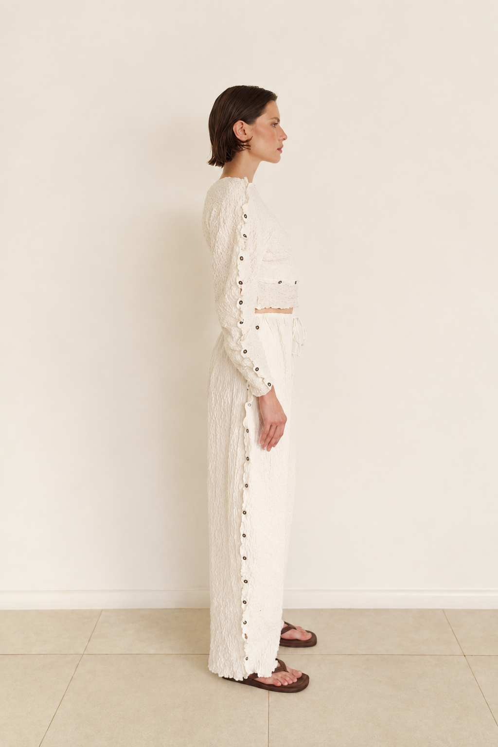 A model stands in profile wearing a long-sleeve white textured knit outfit with decorative dark buttons down the front, paired with brown sandals, against a plain cream wall and light tile floor.