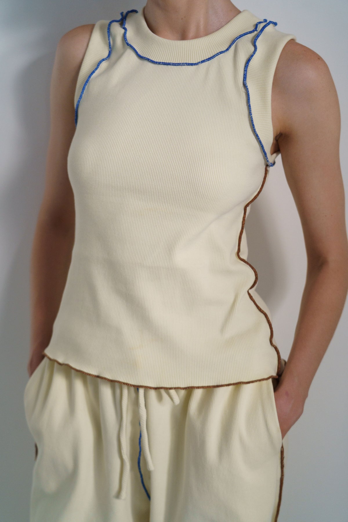A cream-colored sleeveless knit top with blue and brown contrast stitching, shown on a model against a plain gray background.