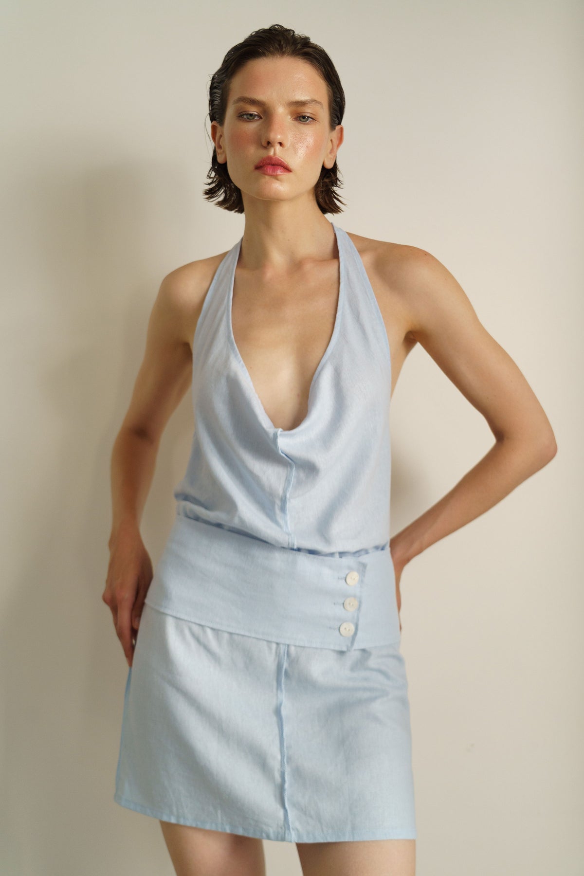 A model wearing a light blue sleeveless halter top and matching high-waisted mini skirt with three front buttons, posed against a plain beige studio background.