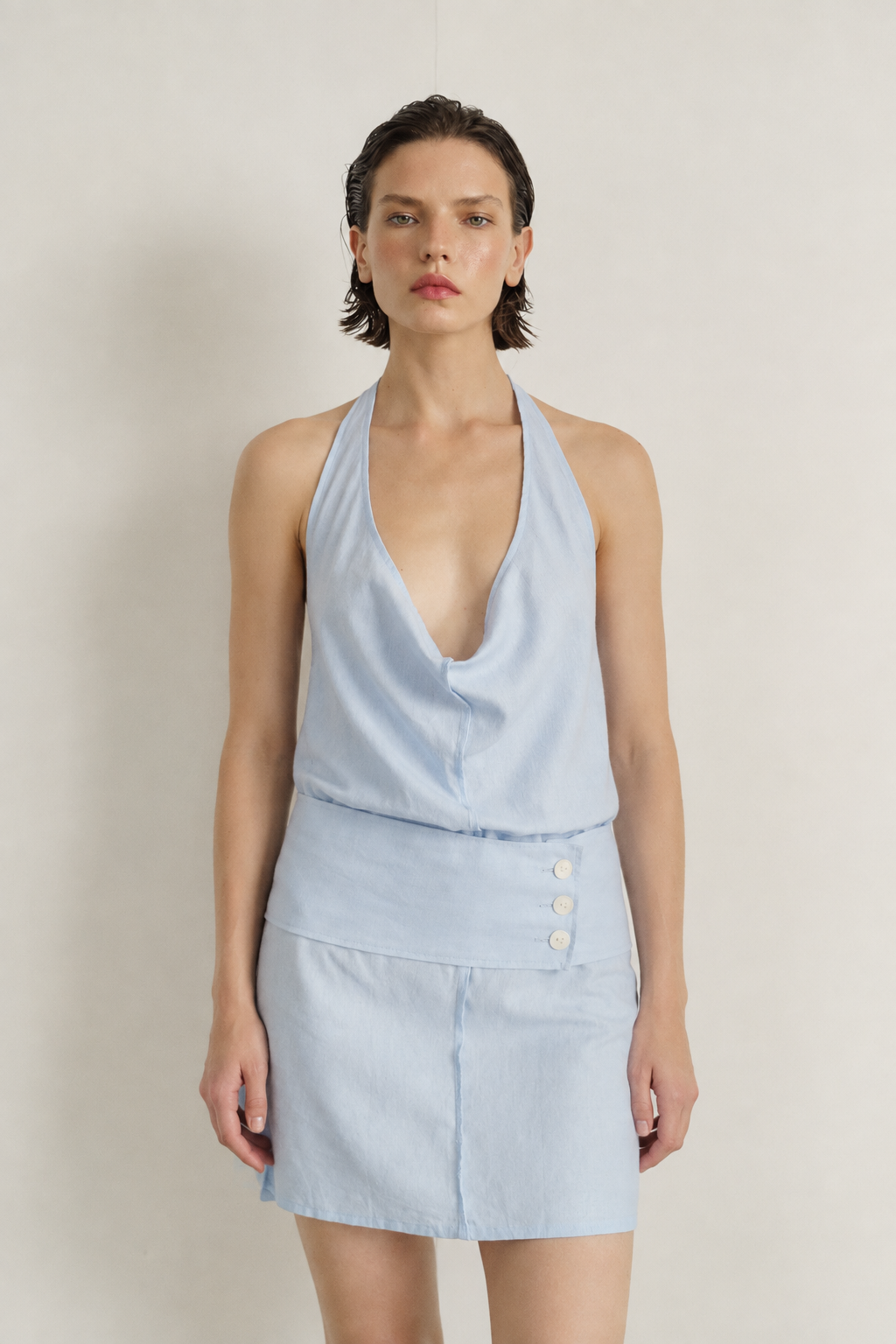 A model wearing a light blue sleeveless halter top and matching high-waisted mini skirt with three front buttons, set against a plain light beige studio background.