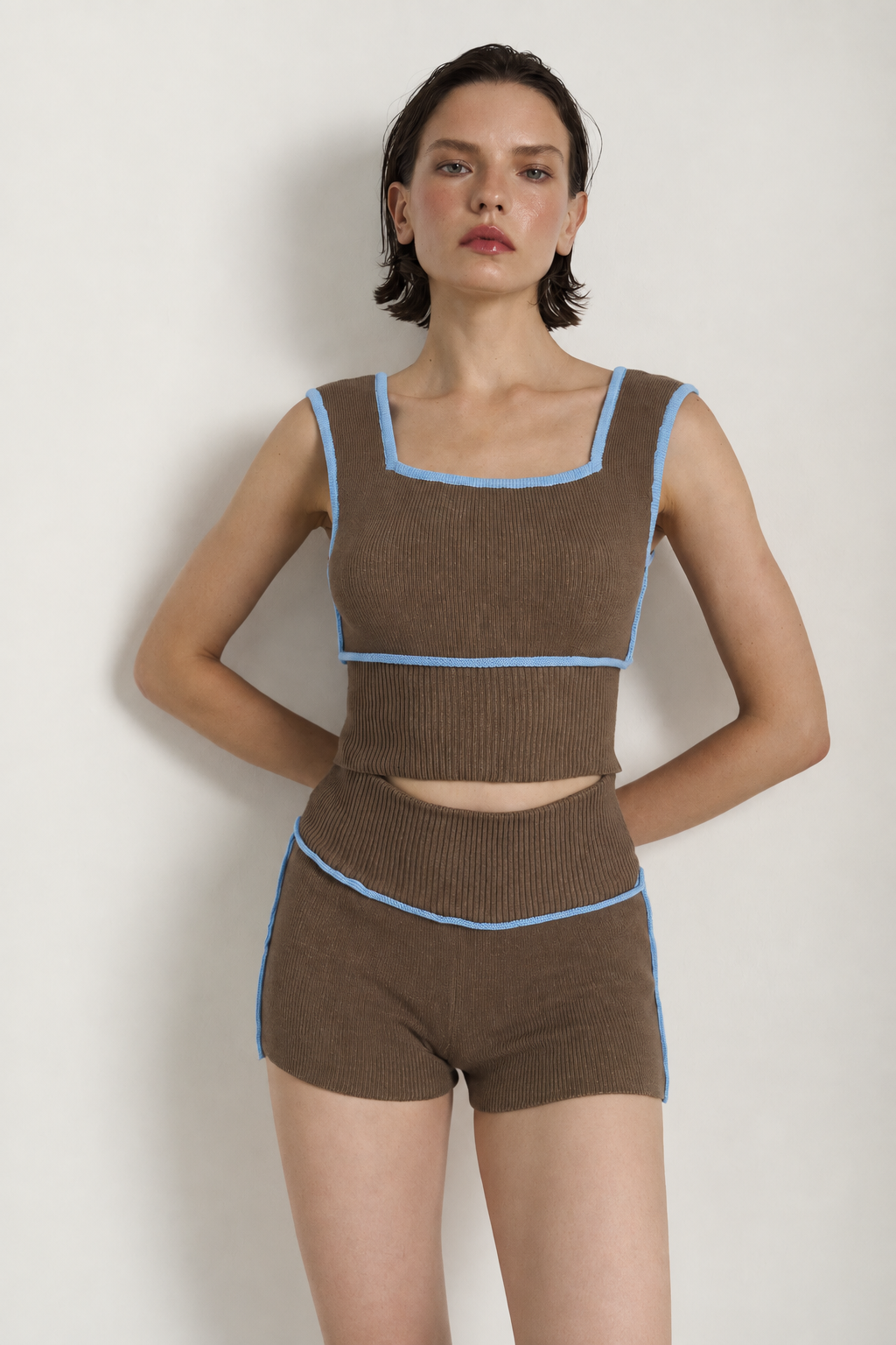 A model wearing a brown ribbed knit two-piece set consisting of a cropped tank top and matching shorts, both featuring light blue trim details, set against a plain off-white background.