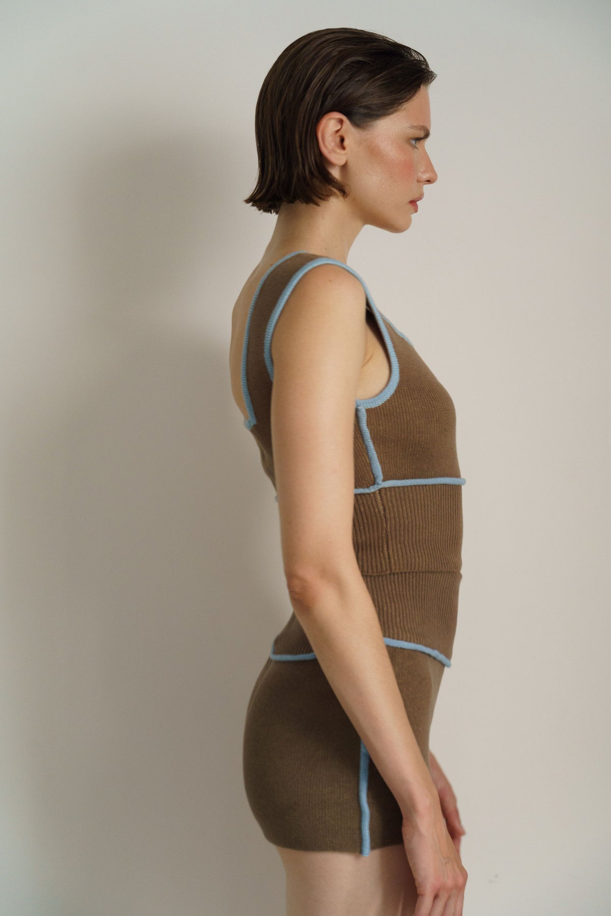 A woman in side profile wearing a brown ribbed knit one-piece bodycon garment with light blue contrast piping, photographed against a plain light gray studio background.