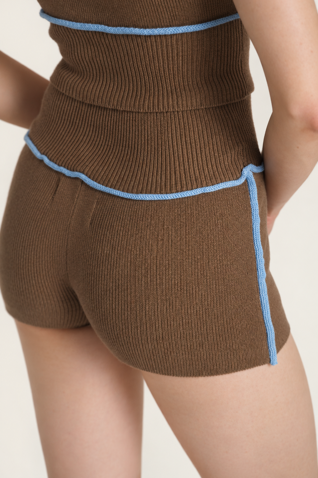 Close-up of a model wearing brown ribbed knit shorts with light blue piping trim, showing the back and side details against a light beige background.