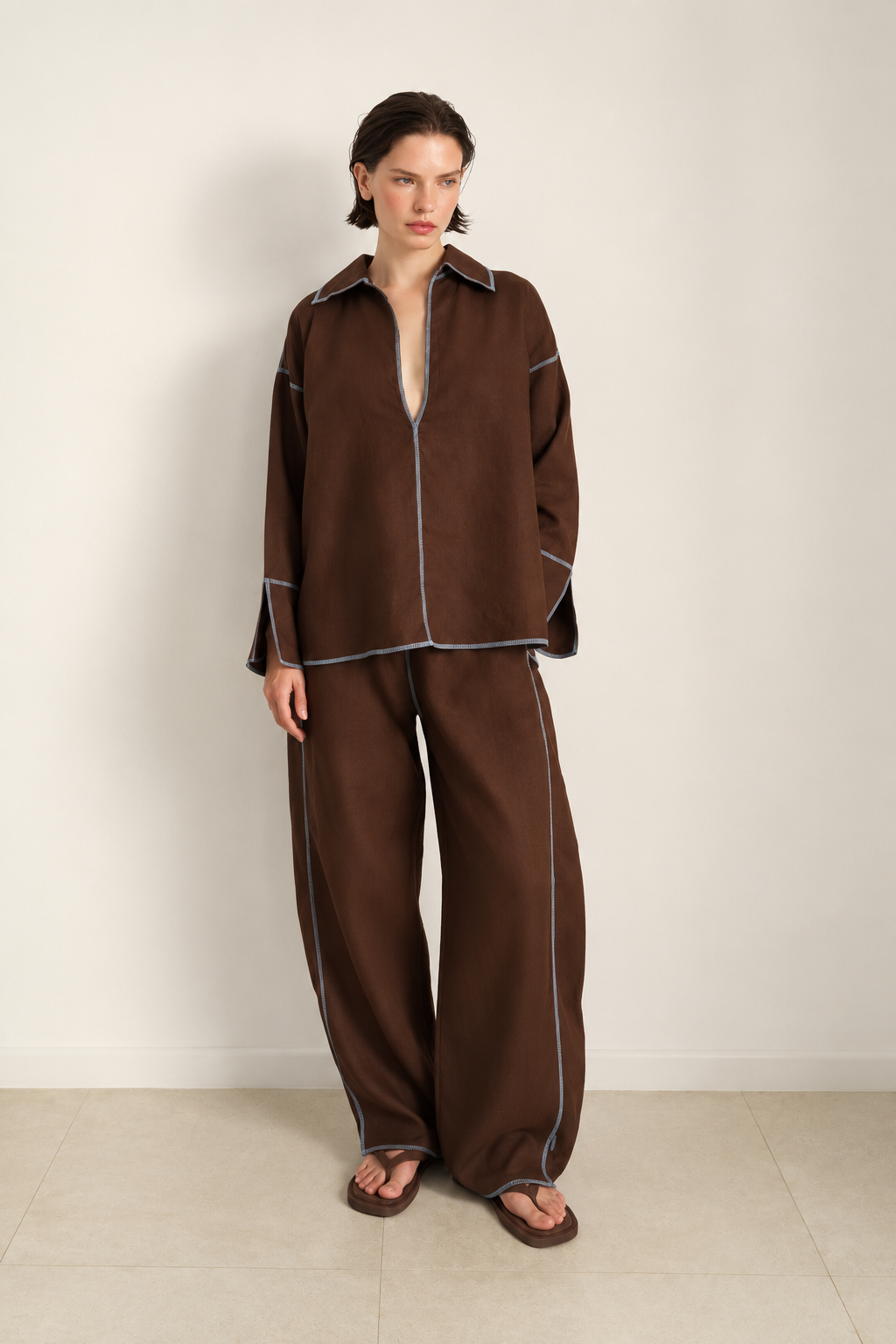 A model stands against a plain off-white wall wearing a matching brown lounge set with light blue piping details, consisting of a long-sleeve collared top and wide-leg pants.