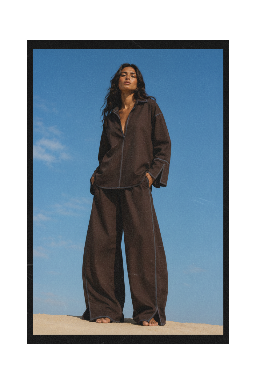 A woman stands outdoors on a sandy surface against a clear blue sky, wearing a dark brown matching lounge set with a button-front long-sleeve top and very wide-leg pants.