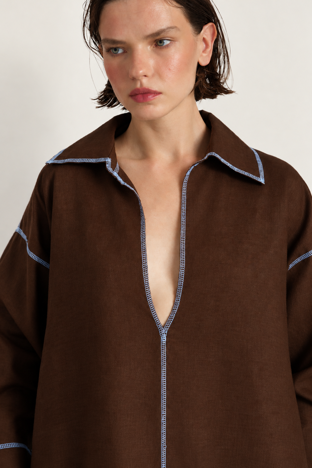 A close-up of a model wearing a brown linen shirt with light blue contrast stitching along the collar, front opening, and cuffs.