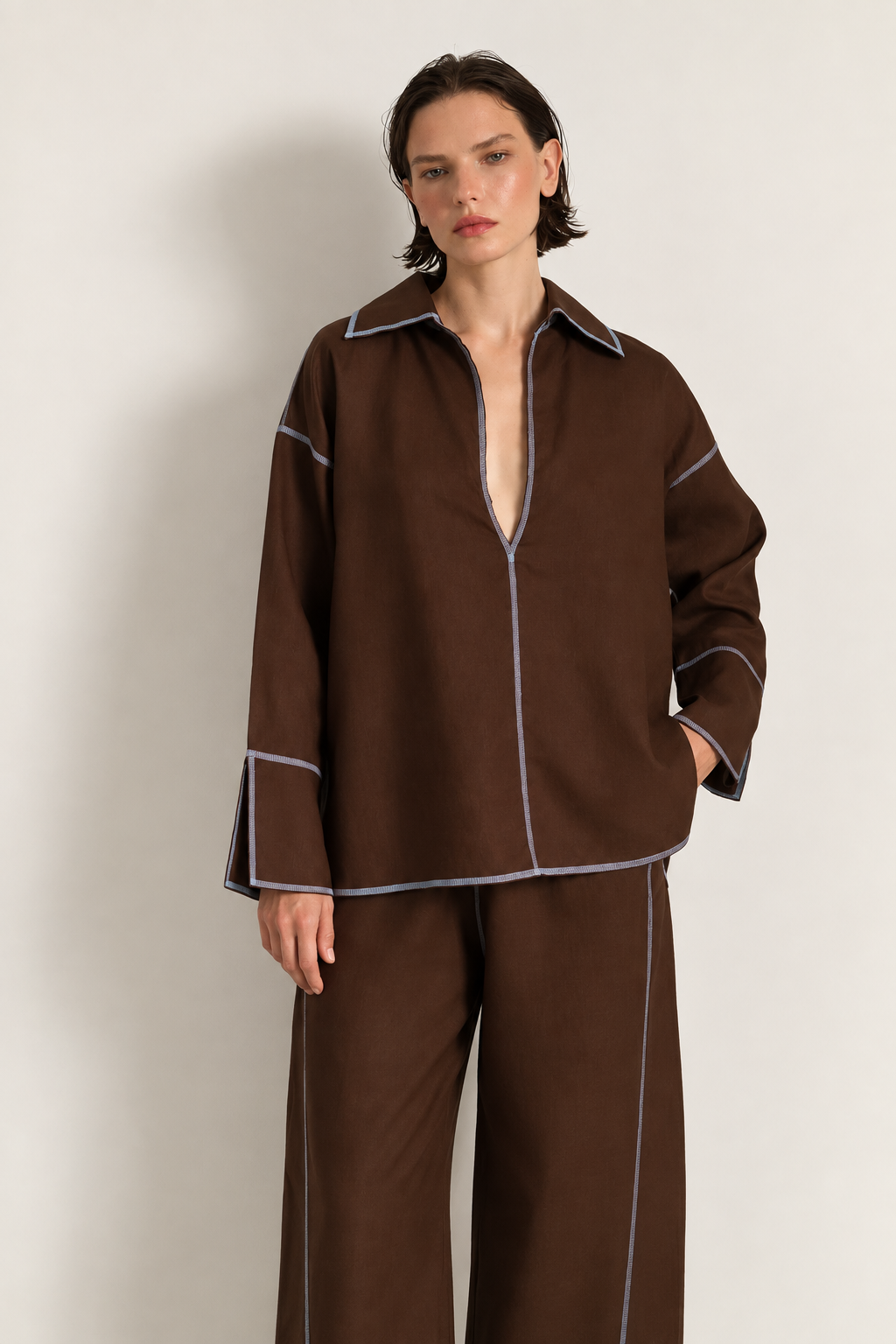 Woman wearing a brown linen set with blue overlock stitching on the seams. She is having her hands in her pockets, staring at the camera.