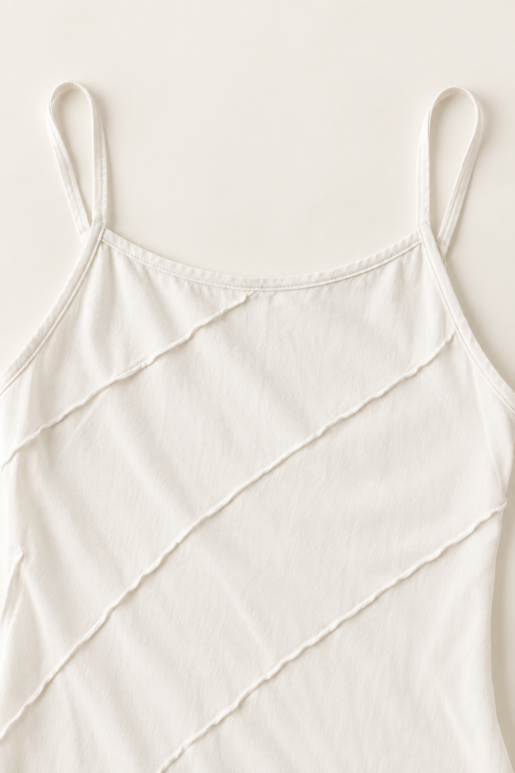A close-up of a light cream or off-white camisole top with thin spaghetti straps and diagonal textured stitched lines, shown against a matching light cream background.