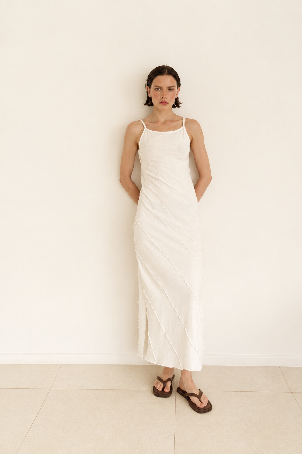 A woman wearing a white spaghetti-strap maxi dress with diagonal textured stitching, standing against a minimalist off-white wall.