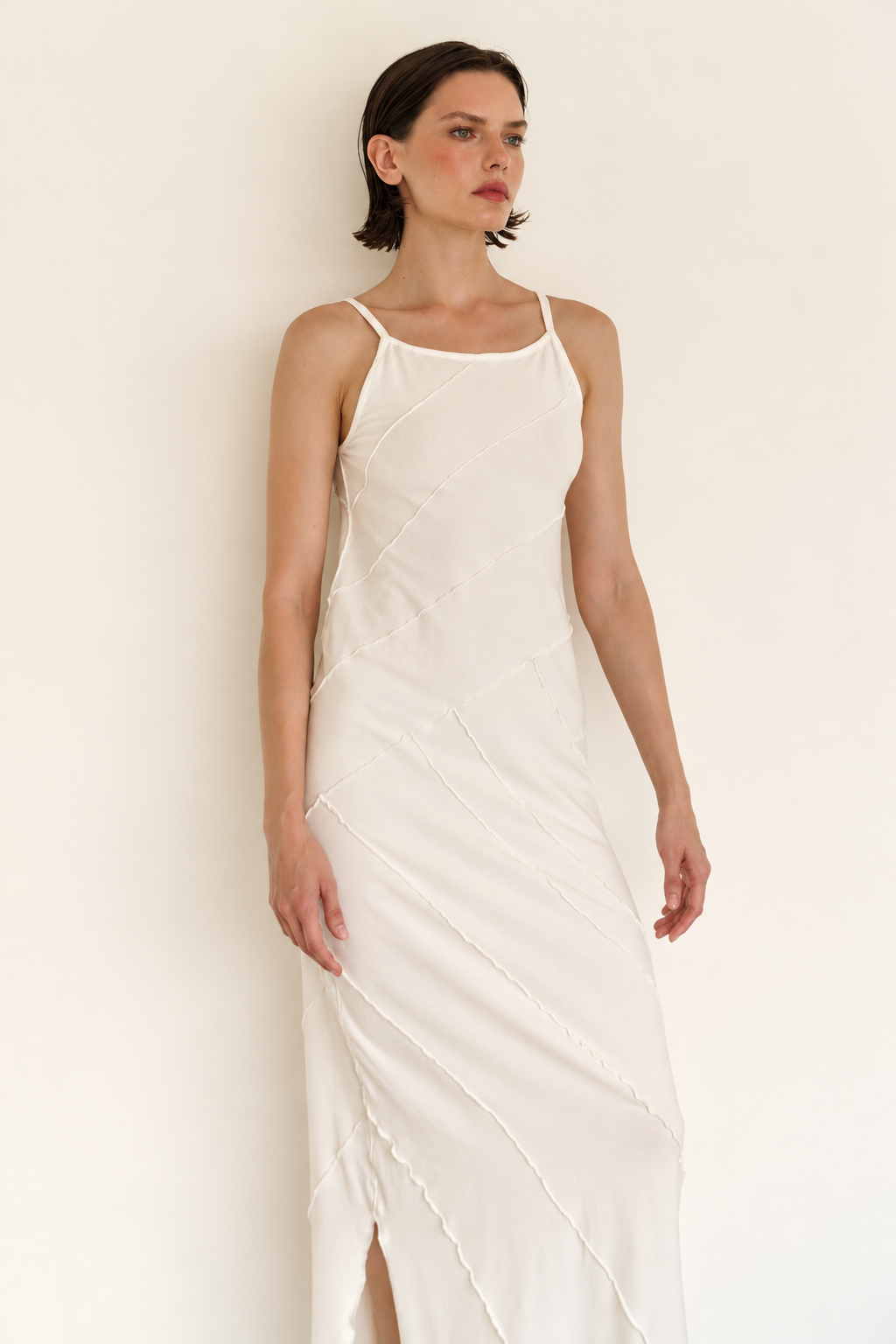 A woman modeling a sleeveless, off-white slip dress with thin straps and diagonal gathered seams, standing against a plain off-white wall.