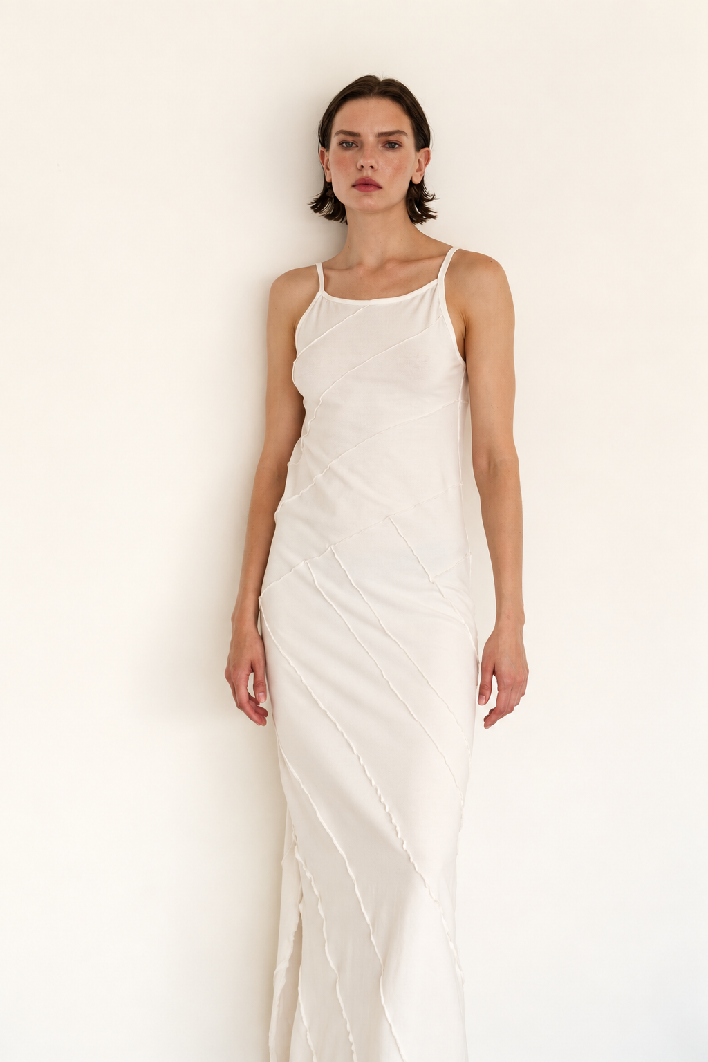 A woman modeling a white sleeveless slip dress with thin straps and a draped, gathered texture, standing against a plain off-white wall.