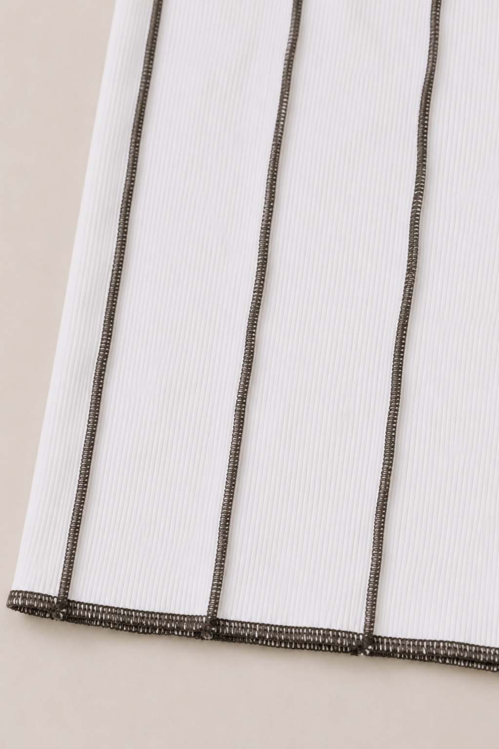 Close-up of a white ribbed fabric with three vertical dark metallic stripes and a matching striped hem, shown on a light neutral background.