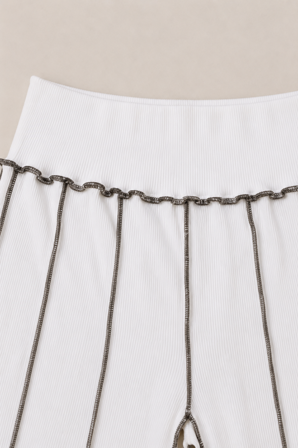 Close-up of a white ribbed garment with dark metallic chain detailing hanging vertically from a waistband, against a light beige background.