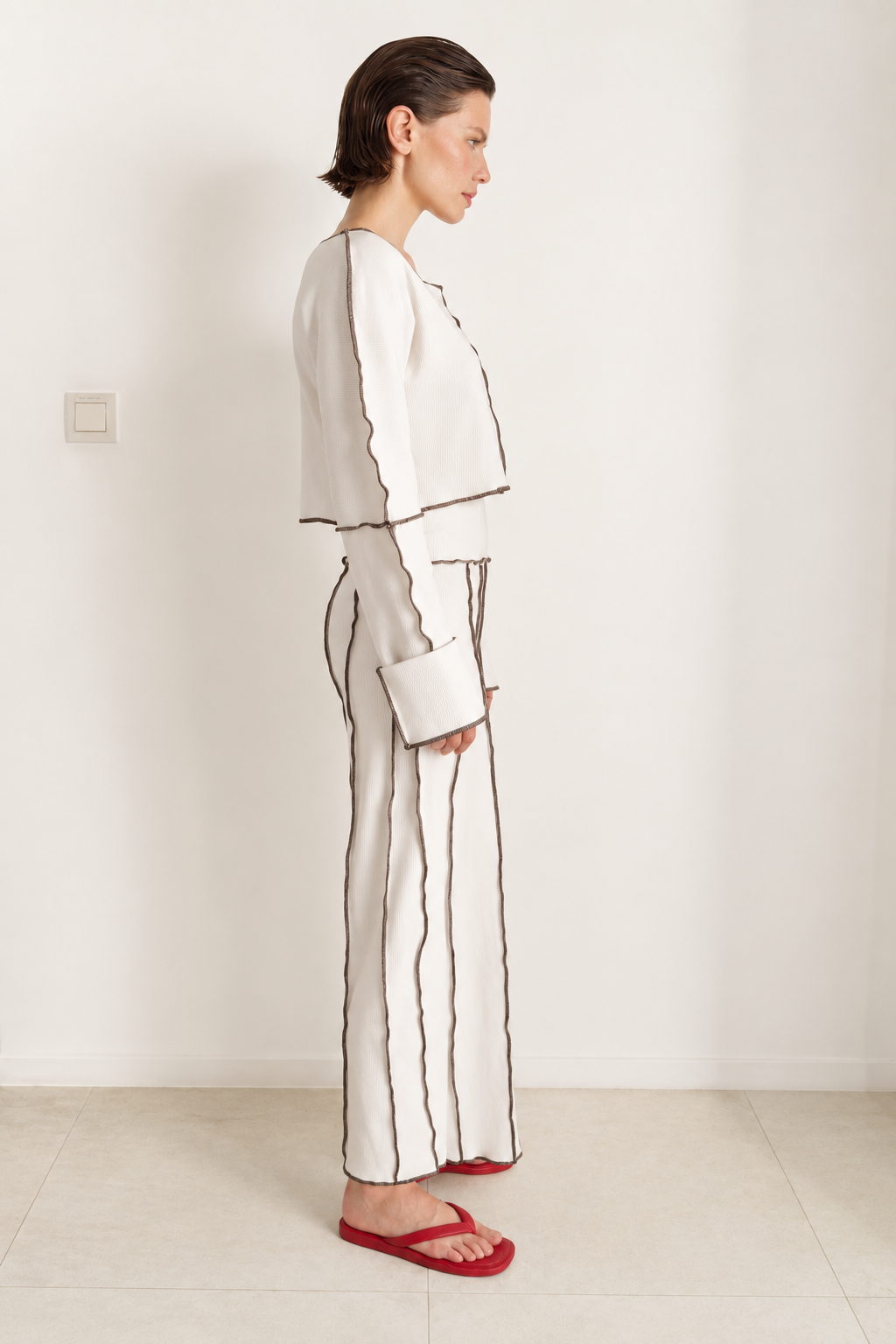 A woman stands in profile wearing a matching off-white two-piece set with dark brown piping, consisting of a cropped long-sleeve top and wide-leg trousers, paired with red sandals.